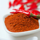 A white dish brimming with vibrant red The Spice Lab Cayenne Pepper X-Hot Ground, crowned by whole red chili peppers, highlights the bold intensity of this fiery seasoning from The Spice Lab, set against a blurred backdrop featuring additional chili peppers.