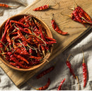 A wooden bowl brimming with dried red chile de arbol peppers sits on a wooden board, with more chiles scattered across a light fabric surface—ideal for grinding into The Spice Lab Chile de Arbol Powder or for stocking up your bulk foodservice pantry with top-quality chiles from The Spice Lab.