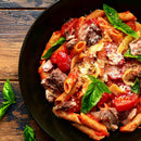 A black plate with penne pasta, cherry tomatoes, shredded meat seasoned with BBQ seasoning, grated cheese, and fresh basil leaves—beautifully garnished with The Spice Lab Savory Whole Leaf 1 lb by The Spice Lab—sits on a wooden table.
