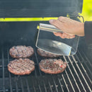 stainless steel burger smasher grill press 5.7 x 6 inches with ergonomic handle shown with burgers on charcoal grill 