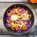 A black bowl filled with shredded purple cabbage, grated carrot, corn, sprouts, and a dollop of hummus—lightly seasoned with The Spice Lab Ground Celery Seeds by The Spice Lab—sits on a wooden table with utensils nearby.