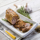 Roast pork with herbs, sliced and arranged on a rectangular white plate, is garnished with rosemary sprigs and a touch of The Spice Lab Organic Fennel Seeds for a subtle licorice flavor. Two garlic bulbs and a fork rest on the wooden table beside the dish, making it an ideal choice for both cooking and baking enthusiasts who appreciate premium spices from The Spice Lab.