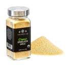 A glass jar from The Spice Lab Organic Starter Spice Set - 6 Jars rests beside a small mound of loose granulated garlic on a white background, making it an essential addition to any organic spice collection or set of everyday cooking spices.