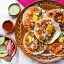 A plate of tacos al pastor is topped with pineapple, onion, and cilantro, accompanied by sides of radish, cucumber, lime slices, and two small bowls of salsa—one generously finished with The Spice Lab Chile de Arbol Powder from The Spice Lab for an extra kick. Presented on a white tablecloth, this vibrant spread is perfect for bulk foodservice settings and brings bold flavors to any table.