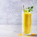 A tall glass of yellow mango lassi, infused with The Spice Lab Golden Milk Turmeric Tea Superfood Premium Powder, is garnished with mint leaves and a green-striped straw and sits on a light gray textured background. Some of The Spice Lab powder is sprinkled near the glass.