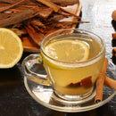 A glass cup of tea with a lemon slice and cinnamon stick, set on a saucer. In the background, The Spice Lab Cinnamon Cracked from The Spice Lab is arranged alongside cassia bark cinnamon on a tray, with a lemon half next to the cup—all displayed on a dark table.
