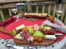 A round patio table with a red placemat holds a charcuterie board of meats, cheeses, apple slices, dried fruit, bread, and Terrapin Ridge Farms Mango Habanero Jam. Two glasses of white wine, a jar of mustard, and napkins are also on the table.