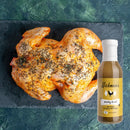 A whole roasted chicken with herbs on a dark slate board, accompanied by a bottle of Hickman's BBQ Marinade Package with Injector from Hickman's BBQ, set against a green textured background.