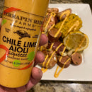 A hand holds a bottle of Terrapin Ridge Farms Chile Lime Aioli Squeeze Garnishing Sauce over seafood boil with corn, potatoes, sausage, and lemon. Perfect for seafood tacos or adding zesty flavor to your next meal.