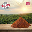 A mound of reddish-brown Rico Rico Chipotle Morita Chile Powder sits on a wooden surface in front of a green field, its smoky, savory flavor promising the bold taste of authentic Mexican cuisine, with the Rico Rico logo and "Made in Mexico" proudly displayed in the sky.