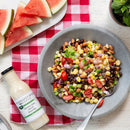 A bowl of bean salad with corn, tomatoes, and onions sits on a gray plate with a spoon. Nearby are slices of watermelon and a quart bottle of Terrapin Ridge Farms Cilantro Lime Ranch Dressing on a red checkered tablecloth.