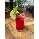 A vibrant pink Margarita infused with Brushfire Farms Bulk Prickly Pear Simple Syrup, served over ice in a tall glass with a chili rim, and garnished with a lime wheel and fresh mint, sits atop a wooden barrel table outdoors.