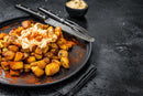 A black plate holds golden-brown roasted potatoes topped with creamy sauce and paprika, seasoned with The Spice Guy Colorado Box blends. A fork and knife sit nearby, with a small bowl of sauce in the background.