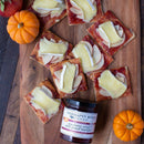 Rectangular crackers with apple slices, melted brie, and Terrapin Ridge Farms Cranberry Relish With Grand Marnier™ are elegantly arranged on a wooden board for holiday entertaining. A jar of the relish sits in front, accented by two small pumpkins.