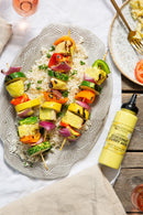Grilled vegetable and pineapple skewers are served over rice on a patterned platter, paired with Terrapin Ridge Farms Coconut Curry Aioli for extra flavor. In the background, a fork rests on a plate of pasta.