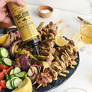A hand drizzles Terrapin Ridge Farms Coconut Curry Aioli over grilled meat and veggie skewers, served with cucumbers, tomatoes, lemon wedges, and red onion. A bowl of the aioli and a glass are in the background.