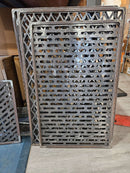 Three DDR Fab Custom Grill Grates (28" x 21" x 1/2") with geometric cutout patterns—two featuring diagonal slots, one with a diamond lattice—lean against a wall. Boxes and the floor appear in the background.