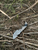 The Texan Knives Exquisite Custom Handmade Damascus Steel Guthook Skinning Knife, featuring a unique resin handle and 7.5” patterned blade with leather sheath, rests outdoors on dry twigs and grass.