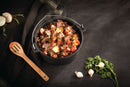 The Napoleon Grills Cast Iron Dutch Oven 4.5qt (56062) in black is filled with beef stew, carrots, and potatoes. It sits on a dark surface beside a wooden spoon, dark cloth, parsley, garlic bulbs, and half an onion.