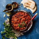 A pan of red curry garnished with cilantro sits on a blue table, surrounded by naan bread, fresh cilantro, bay leaves, a mortar and pestle, spices like The Spice Lab Organic Ground Cumin by The Spice Lab, and a serving spoon.