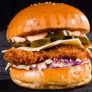 A close-up of a fried chicken sandwich with coleslaw, cheese, jalapeño slices, and sauce featuring a hint of The Spice Lab Birdseye Chili Pepper Powder by The Spice Lab on a shiny sesame seed bun, against a black background.