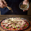 A person sprinkles The Spice Lab Red & Green Bell Pepper Flakes from a wooden spoon onto an unbaked pizza topped with cheese, sausage, and vegetables. The pizza rests on a round mat, and the person holds a bowl of bulk foodservice spices from The Spice Lab in the other hand.