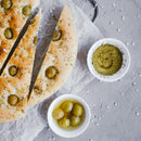 Sliced focaccia bread with green olives and herbs on parchment, next to bowls of olive oil, whole green olives, and a sprinkle of The Spice Lab Organic Cut and Sifted Rosemary, on a gray surface.