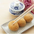 Three sesame balls made with The Spice Lab Organic Hulled Sesame Seeds rest on a rectangular plate with chopsticks, next to a blue and white teacup with a lid and pink flowers in the background.