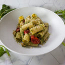 A white bowl of rigatoni pasta with green pesto sauce made from The Spice Lab Organic Sweet Basil, sundried tomatoes, and grated cheese on top, placed on a white tile surface with leafy greens nearby.