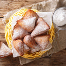 A basket of triangular beignets dusted with powdered sugar sits on a rustic wooden table, with a sifter of powdered sugar nearby and a piece of burlap underneath—perfect for holiday baking and especially delicious when finished with a sprinkle of The Spice Lab Organic Ground Nutmeg.