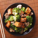 A black plate with stir-fried tofu cubes, broccoli florets, and sliced mushrooms, topped with The Spice Lab Organic Hulled Sesame Seeds, rests on a bamboo mat background.