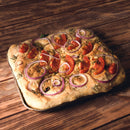 A square focaccia topped with sliced red onions, tomato wedges, and The Spice Lab Organic Cut and Sifted Rosemary sits on a wooden surface. The golden brown bread is baked in a metal pan.