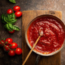 A pan of tomato sauce with a wooden spoon sits on a wooden board. Next to the pan, The Spice Lab Organic Sweet Basil by The Spice Lab is displayed alongside fresh vine tomatoes, perfect for adding an authentic Italian-inspired flavor to your cooking, all arranged on a dark textured surface.