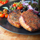 Two grilled pork chops on a black plate, garnished with fresh herbs, cherry tomatoes, red onion slices, and leafy greens. The pork chops feature a browned, seasoned crust that’s elevated with The Spice Lab Ground Black Pepper by The Spice Lab for an extra burst of everyday flavor.