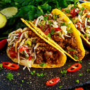 Three crunchy tacos filled with seasoned ground beef, shredded cheese, diced tomatoes, avocado, and fresh herbs are arranged on a dark surface with sliced red chili peppers and cilantro in the background. A sprinkle of The Spice Lab Scotch Bonnet Sea Salt by The Spice Lab adds a bold Caribbean-inspired flavor, perfectly complementing the dish with its spicy finishing salt.