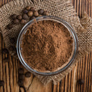 A glass bowl filled with The Spice Lab Organic Ground Allspice sits on a piece of burlap, surrounded by whole allspice berries on a rustic wooden surface—an essential spice from The Spice Lab for crafting authentic Jamaican jerk seasoning.