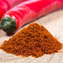 A small mound of The Spice Lab Organic Cayenne Pepper rests on a wooden surface, with two whole red chili peppers in the background, capturing the vibrant essence often found in Indian cooking.