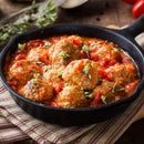 Meatballs in tomato sauce, garnished with The Spice Lab Organic Oregano Leaf, are served in a black skillet and placed on a striped cloth, with herbs and tomatoes in the background—a beautiful nod to classic Italian cooking.
