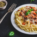 A plate of spaghetti topped with meat sauce, grated cheese, and fresh basil, finished with a sprinkle of The Spice Lab Organic Oregano Leaf—a classic nod to Italian cooking—sits on a dark surface alongside a fork and a small bowl of herbs.