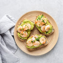 Three slices of bread topped with mashed avocado, cooked shrimp, and a sprinkle of The Spice Lab Whole Leaf Cilantro by The Spice Lab are arranged on a wooden plate, placed on a light gray surface with a folded gray cloth napkin beside the plate.