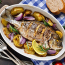 Grilled whole fish on roasted potatoes and red onions, garnished with rosemary and a lemon wedge, served in a white dish on a blue checkered cloth with slices of bread and cutlery nearby—a delicious example of French & Italian-inspired cooking, beautifully elevated by The Spice Lab Flavors of Provence Seasoning Set.