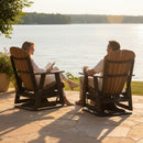 A woman reads and smiles at a man sipping from his mug as they relax by the lake on Kanyon Living Adirondack Poly Glider Swivel Chair Deluxe in Antique Mahogany and Black, enjoying the sun amid trees and water.