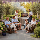 A group relaxes and chats in a garden patio on Kanyon Living Adirondack Poly Glider Swivel Chairs in Light Grey & Weathered Wood, enjoying outdoor comfort as they watch a man grill food on a sunny day, surrounded by flowers.