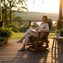 At sunset, a woman relaxes with a mug and book on the Kanyon Living Adirondack Poly Glider Swivel Chair in Black & Deluxe Antique Mahogany. She sits on a cozy deck with potted plants, string lights, and views of lush greenery.