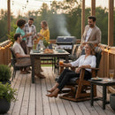 Six adults gather casually outdoors on a wooden deck with string lights, food, and drinks. Some chat near the grill while one woman relaxes in a Kanyon Living Adirondack Poly Glider Swivel Chair in Black & Deluxe Antique Mahogany.