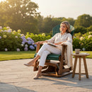 A woman relaxes with a book on the Kanyon Living Adirondack Poly Glider Swivel Chair in Dark Green & Cedar. Dressed in light clothes, she enjoys a sunny patio with hydrangeas and greenery, a blanket draped over the chair, and a cup beside her.