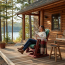 A woman in a white sweater and boots relaxes with her mug on the porch of a log cabin, seated on the Kanyon Living Adirondack Poly Glider Swivel Chair (Dark Green & Cherry). A blanket and cup are nearby, with trees and a lake softly shining in the background.