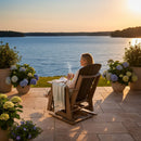 At sunset, a woman relaxes on a Kanyon Living Adirondack Poly Glider Swivel Chair Deluxe in Brazilian Walnut & Weathered Wood, holding a cup and facing the lake. Potted hydrangeas, a blanket, and an open book complete her lakeside patio escape.