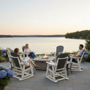 Four people relax in Kanyon Living Adirondack Poly Glider Swivel Chairs (Driftwood Grey & White) around a fire pit on a lakefront patio at sunset, with blue hydrangeas and tranquil water and trees enhancing the serene outdoor setting.