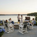 At sunset, three people relax in Kanyon Living Adirondack Poly Glider Swivel Chairs in Driftwood Grey & White, while two stand by the grill on a lakeside patio with blue hydrangeas and a fire pit, enjoying a peaceful evening.